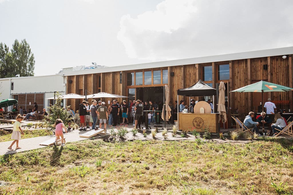 shapers-club- A group of people are gathered outside a wooden building under several umbrellas. Some are standing, while others are seated at tables. Nearby, children are playing on the grass. The sky is partly cloudy, and the atmosphere appears relaxed and social. -surf