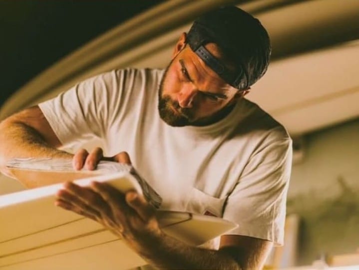 shapers-club- A bearded man wearing a cap and a white t-shirt carefully shaping a piece of material, possibly a surfboard, with a focused expression. The background is softly lit, highlighting the man's concentration and attention to detail. -surf