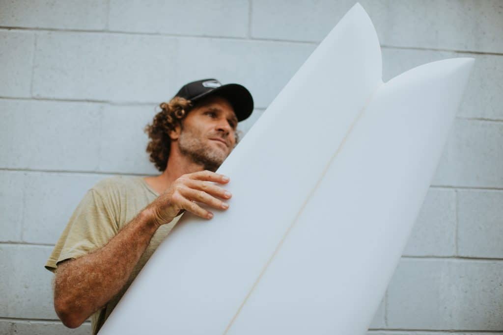 shapers-club- Un homme aux cheveux frisés et à la casquette tient une planche de surf blanche à la verticale contre un mur de briques gris clair, perdu dans les pensées automobiles de Brouillon. -surf