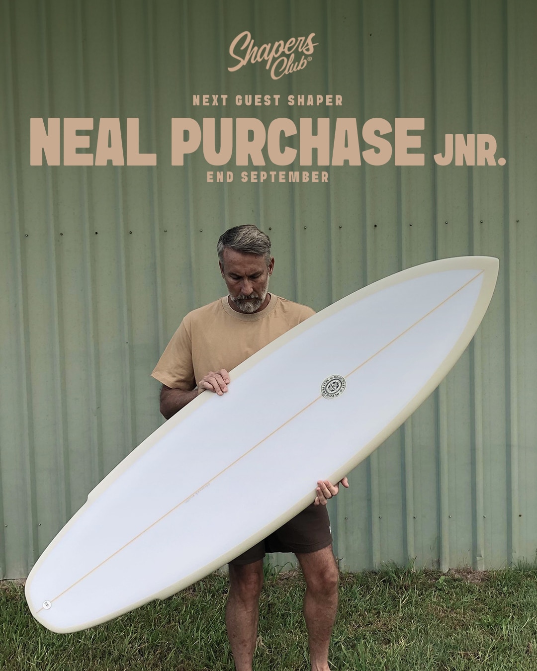 shapers-club- Un homme se tient à l'extérieur contre un mur en tôle ondulée, tenant une planche de surf blanche. Le texte au-dessus indique : "Le Shapers Club accueille Neal Purchase Jnr. comme prochain shaper invité. Fin septembre". -surf