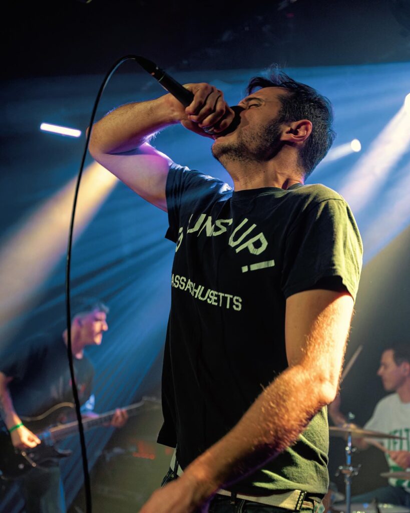 shapers-club- Un homme chante passionnément dans un micro sur la scène du Shapers Club House, vêtu d'un t-shirt noir, tandis qu'un groupe joue de la guitare et de la batterie en arrière-plan sous des lumières de scène colorées, capturant ainsi l'énergie brute des concerts. -surf