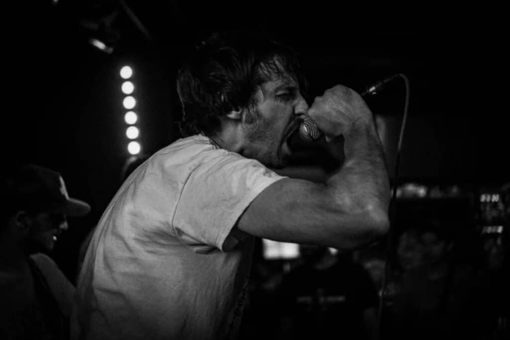 shapers-club- Un homme hurle passionnément dans un microphone lors d'un concert de HARDCORE SESSION au Shapers Club House, avec un autre musicien en arrière-plan. L'image en noir et blanc capture l'émotion intense et l'énergie brute typiques des concerts underground. -surf