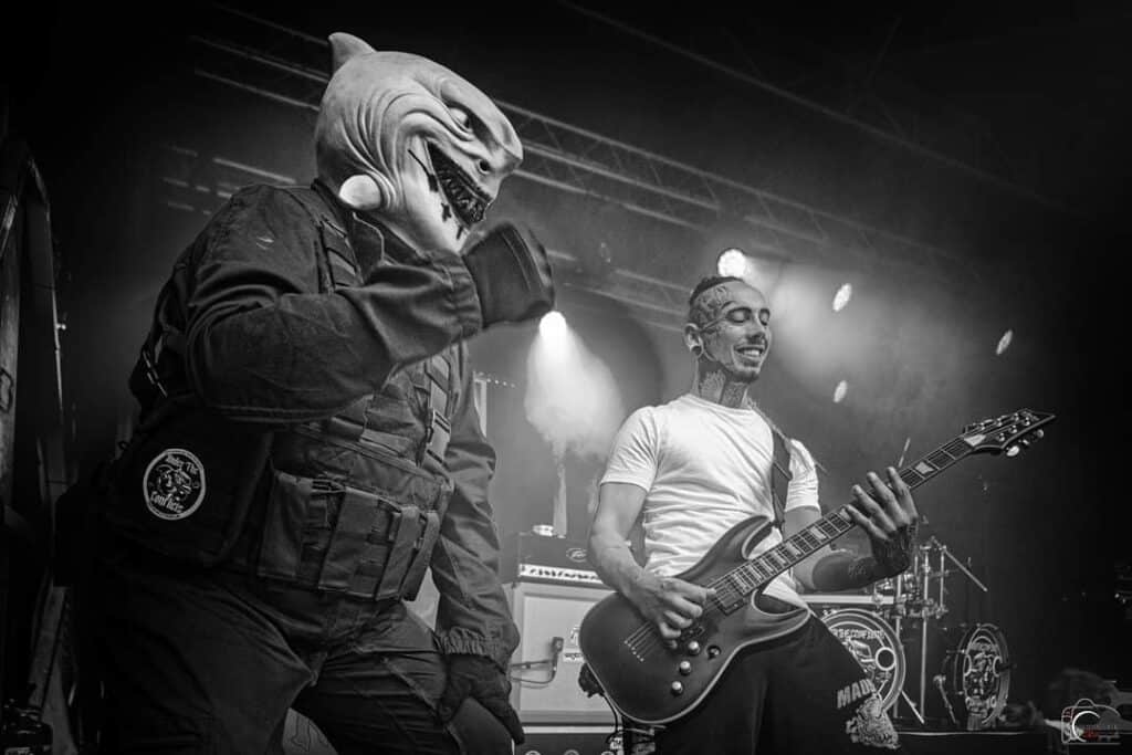 shapers-club- Un musicien portant un masque de requin et un équipement tactique monte sur scène aux côtés d'un guitariste tatoué vêtu d'un t-shirt blanc. Les lumières de la scène s'allument derrière eux, capturant l'énergie brute d'un concert de HARDCORE SESSION en noir et blanc. -surf