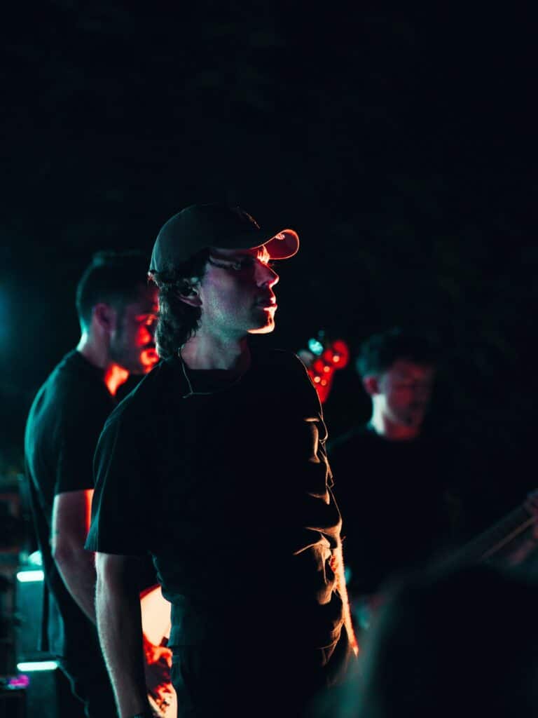 shapers-club- Un homme portant une casquette de base-ball et une chemise sombre se tient sous un éclairage de scène spectaculaire au Shapers Club House, avec deux autres personnes légèrement floues derrière lui, ce qui suggère une performance musicale en direct ou l'un de leurs concerts de session hardcore. -surf