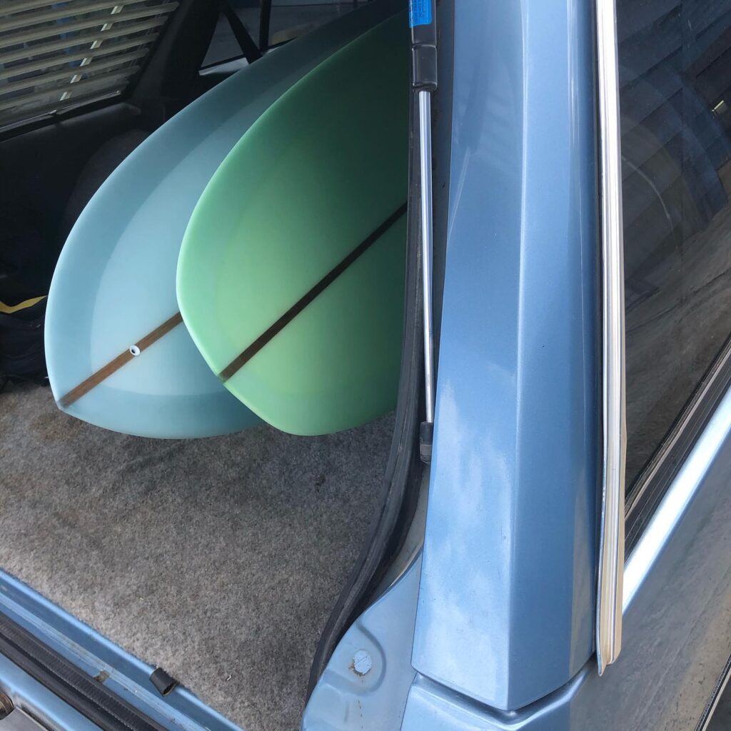 shapers-club- Deux planches de surf, l'une bleu clair et l'autre vert clair, reposent sur une moquette beige à l'arrière d'une voiture bleue dont le hayon est ouvert. Une scène qui rappelle un road trip de Champion du Monde ou une aventure de la Résidence de Shape. -surf