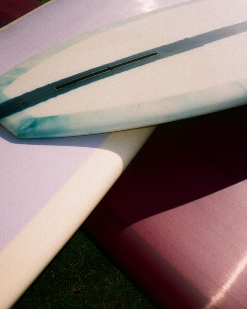 shapers-club- Gros plan sur trois planches de surf du Shapers Club qui se chevauchent sur l'herbe, montrant leurs surfaces texturées et leurs couleurs douces - rose, crème et blanc avec une bande sarcelle - clin d'œil au savoir-faire artisanal de chaque résidence de shape. -surf