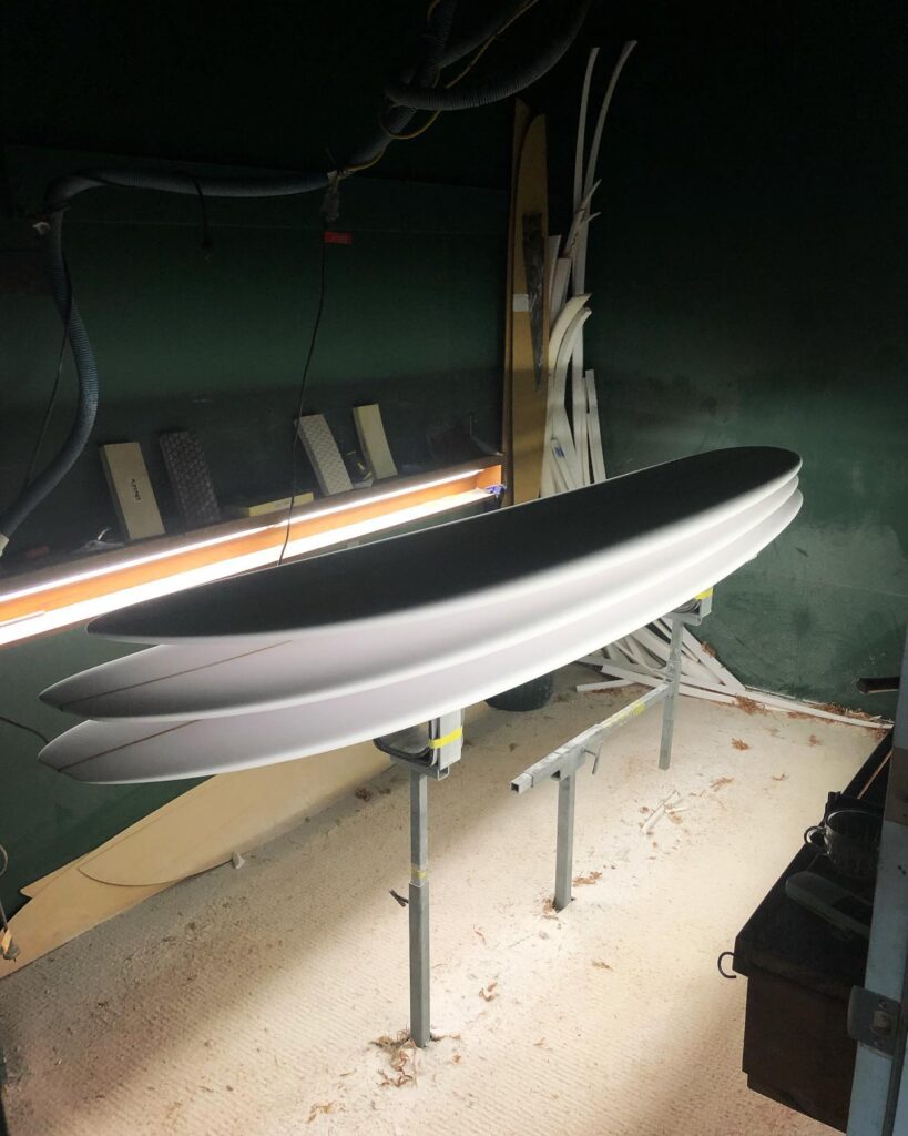 shapers-club- Une planche de surf fraîchement façonnée repose sur des supports dans un atelier faiblement éclairé du Shapers Club, avec un éclairage fluorescent mettant en valeur ses contours blancs et lisses et d'autres planches de surf empilées à l'arrière-plan. -surf