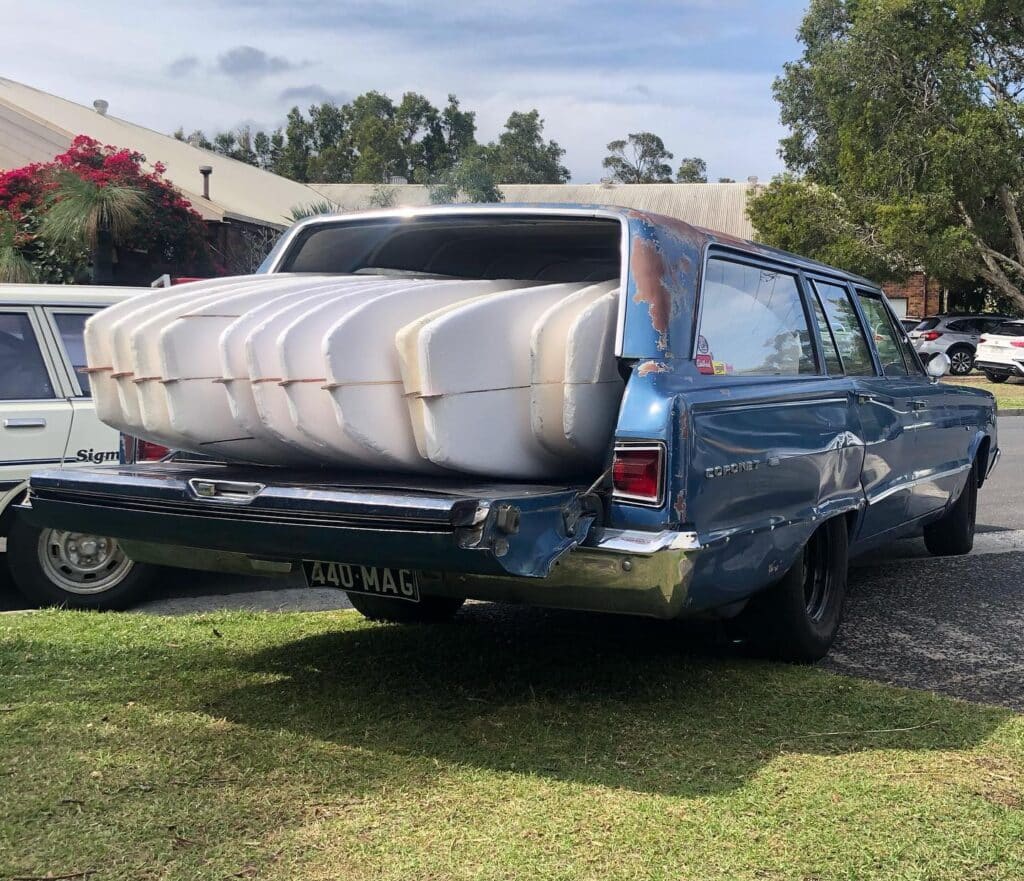 shapers-club- Un break bleu d'époque à l'extérieur rouillé est garé avec le hayon arrière ouvert, transportant plusieurs grandes planches de surf blanches du Shapers Club qui dépassent de l'arrière. La voiture est sur l'herbe, à côté d'un véhicule blanc et de quelques arbres. -surf