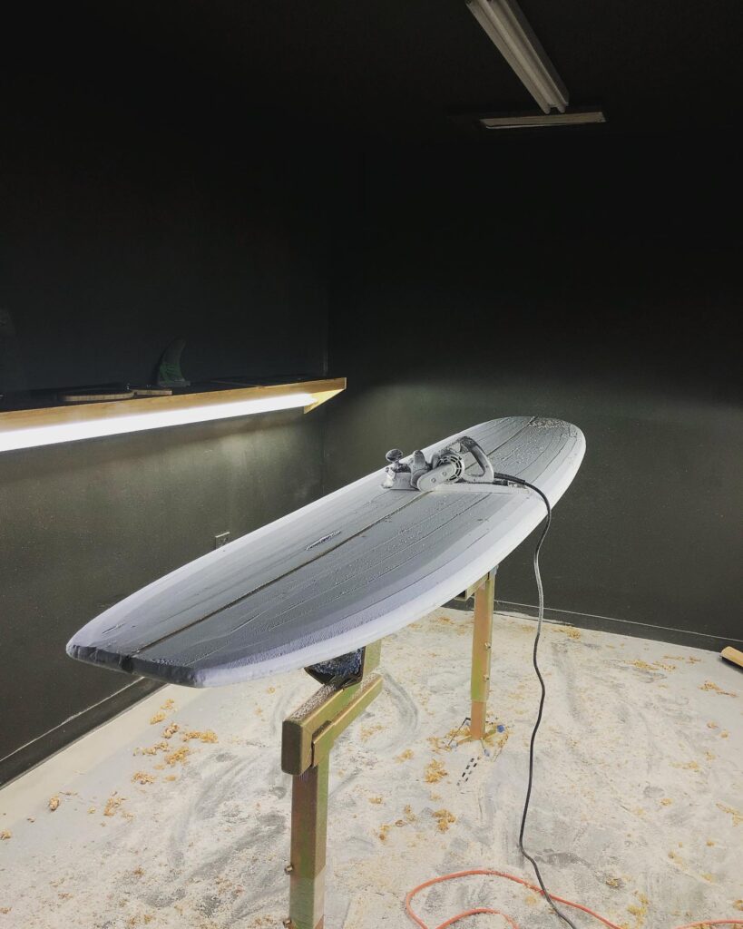 shapers-club- Une planche de surf est montée sur un support dans un atelier du Shapers Club, partiellement façonnée avec un outil électrique posé sur le dessus. Des copeaux de mousse recouvrent le sol et des lampes de plafond éclairent l'espace de travail créatif de Kai Ellice-Flint sur des murs sombres. -surf
