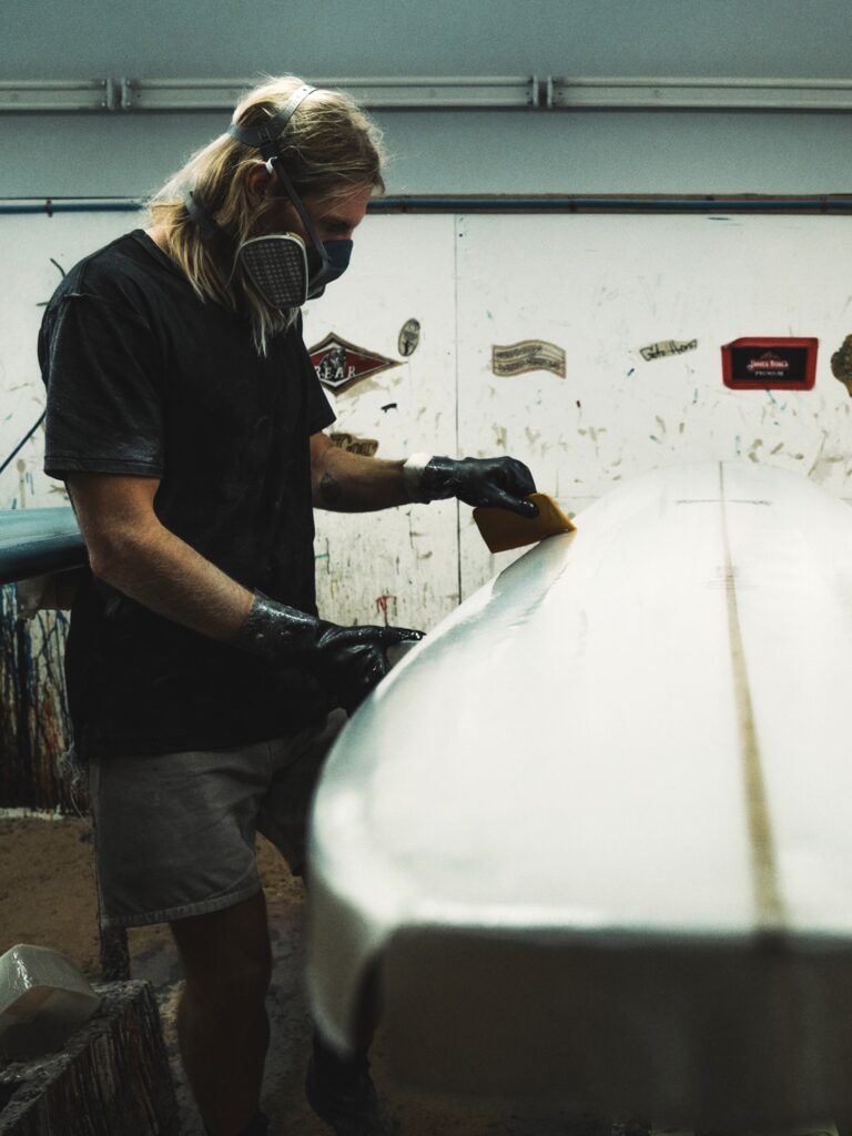shapers-club- Une personne portant des gants, un masque et des protections auditives ponce une planche de surf au Shapers Club, un atelier dont les murs sont couverts de peinture et d'autocollants liés au surf. -surf