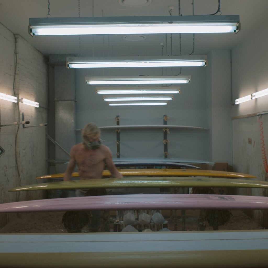shapers-club- Une personne torse nu aux longs cheveux blonds se tient dans un atelier du Shapers Club, entourée de planches de surf de différentes couleurs sous des lumières fluorescentes. Les planches de surf sont empilées sur des étagères le long du mur du fond. -surf