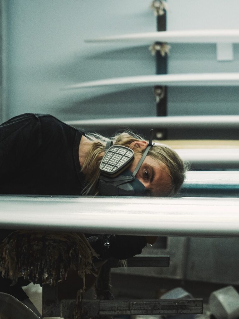 shapers-club- Une personne aux longs cheveux blonds, portant une chemise noire et un masque respiratoire, se penche de près sur une planche de surf dans l'atelier du shapers club, avec plusieurs planches de surf empilées à l'arrière-plan. -surf