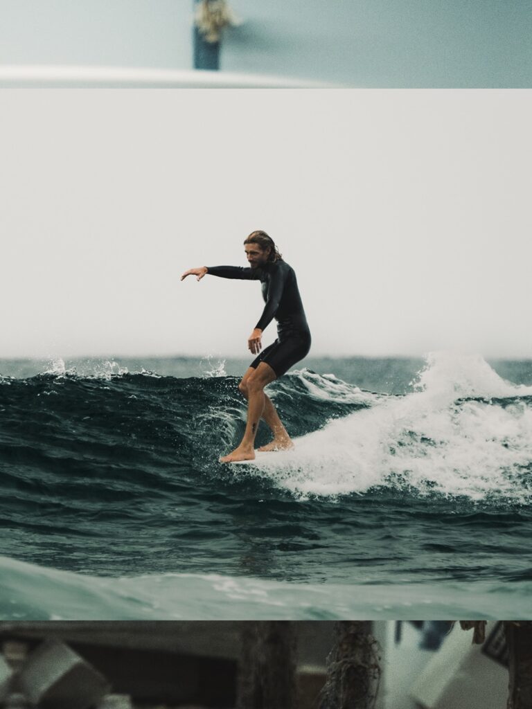 shapers-club- Une personne vêtue d'une combinaison de plongée noire, peut-être du Shapers Club, surfe sur une vague de l'océan, en équilibre avec les bras tendus. Le ciel est couvert et l'eau est sombre et agitée. -surf