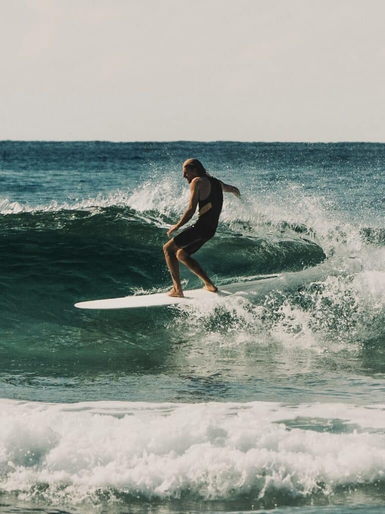 shapers-club- Un surfeur en combinaison noire chevauche une vague sur une planche de surf blanche, entouré par l'eau bleue de l'océan et l'écume blanche sous un ciel lumineux - une scène inspirée par l'art de Kai Ellice-Flint du Shapers Club lors de sa résidence de shape. -surf