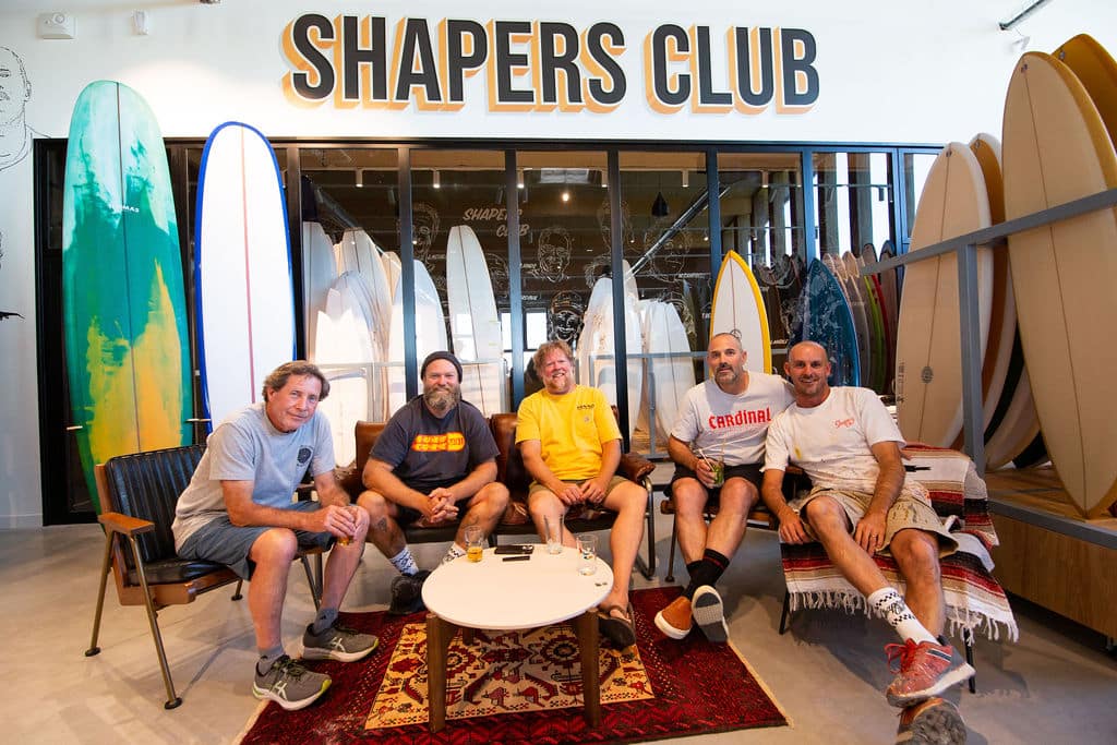 shapers-club- Cinq hommes sont assis sur des chaises et un canapé devant des planches de surf au Shapers Club. Ils sont souriants et détendus, avec des planches de surf exposées sur des étagères derrière eux et un tapis et une table basse au premier plan. -surf