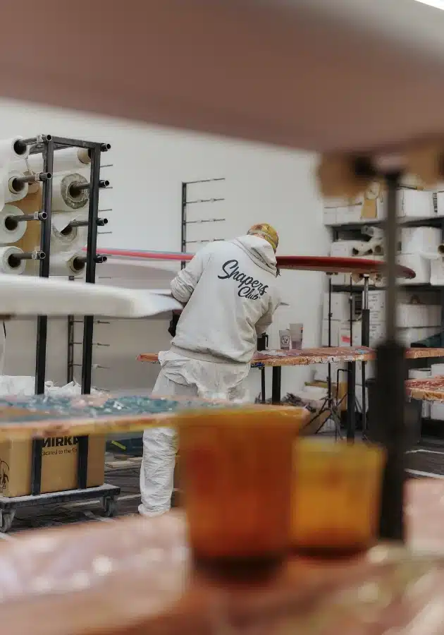 shapers-club- Une personne en combinaison blanche travaille sur une planche de surf dans un atelier, entouré de rayonnages contenant des rouleaux de matériel, d'étagères contenant des fournitures et d'une boîte en carton posée sur le sol. -surf